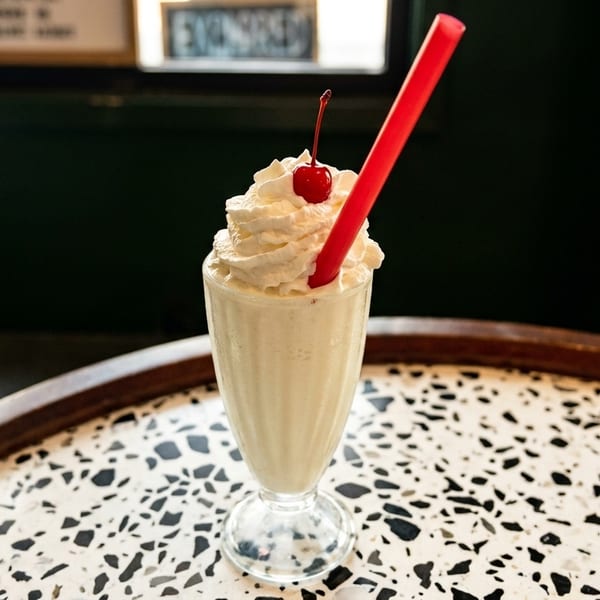 Vanilla milkshake with whipped cream, a cherry, and a red straw on a speckled table.